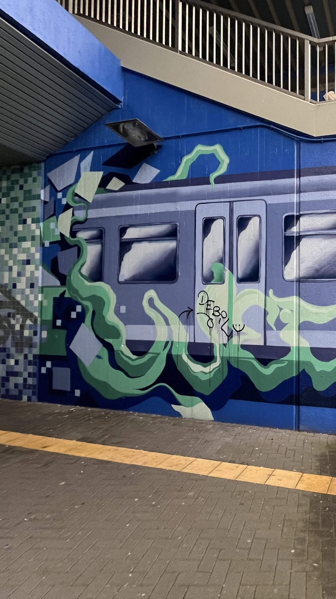 Graffiti eines blauen Zuges an einer Wand unter einer Brücke. Um den Zug herum schlängeln sich grüne, nebelartige Formen. Im Hintergrund Kacheln in Blautönen und eine Geländerkonstruktion.