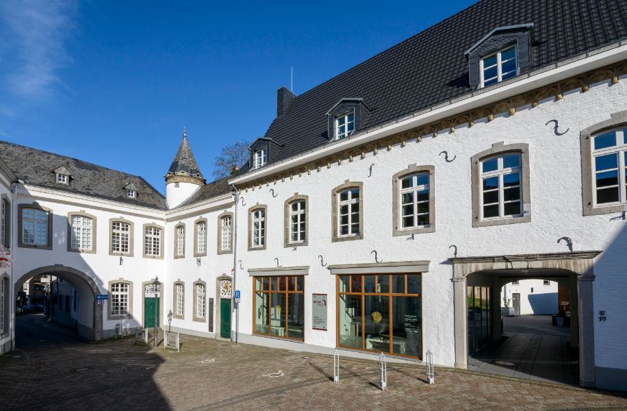 Ein weißes historisches Gebäude mit schwarzen Dachziegeln, Bogenfenstern und einem kleinen Türmchen in einer gepflasterten Fußgängerzone unter strahlend blauem Himmel.